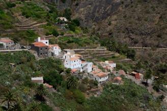 A small, lush rural village nestled within terraced hills and dense vegetation. The village is composed of white and earthy-toned buildings with red-tiled roofs. The landscape surrounding the village is characterized by steep, rocky hillsides and a variety of greenery, including palm trees and other shrubs.