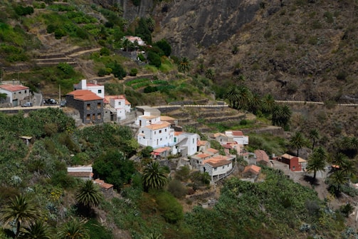 A small, lush rural village nestled within terraced hills and dense vegetation. The village is composed of white and earthy-toned buildings with red-tiled roofs. The landscape surrounding the village is characterized by steep, rocky hillsides and a variety of greenery, including palm trees and other shrubs.