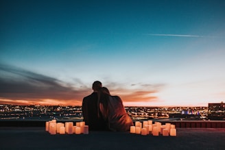 A couple sits closely together on a rooftop overlooking a cityscape during sunset. The sky is filled with gradient hues of orange, blue, and purple. Numerous candles are arranged around them, casting a warm glow.