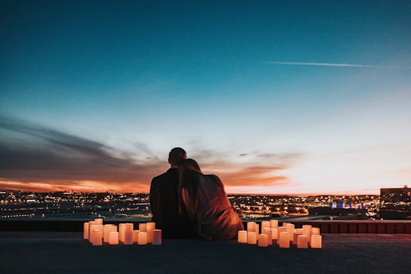 A couple sits closely together on a rooftop overlooking a cityscape during sunset. The sky is filled with gradient hues of orange, blue, and purple. Numerous candles are arranged around them, casting a warm glow.