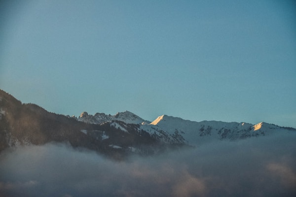 A serene mountain landscape with snow-covered peaks illuminated by soft golden sunlight. Mist gently rolls over the dark forested foothills, under a clear, pale blue sky.