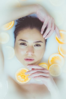 A person is submerged in a milky liquid with several orange and lemon slices floating around. The person's face is partially above the surface, with closed lips and calm expression. Their hands are gently placed near their face.