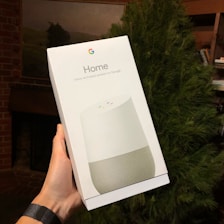 A hand is holding a boxed product labeled 'Home' with the Google logo, indicating it's a voice-activated speaker by Google. The background includes a partially visible tree and a room setting with a fireplace.