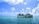 A catamaran is anchored on a clear turquoise sea under a bright blue sky with scattered white clouds. The vessel is surrounded by calm waters, and a distant island is visible on the horizon.