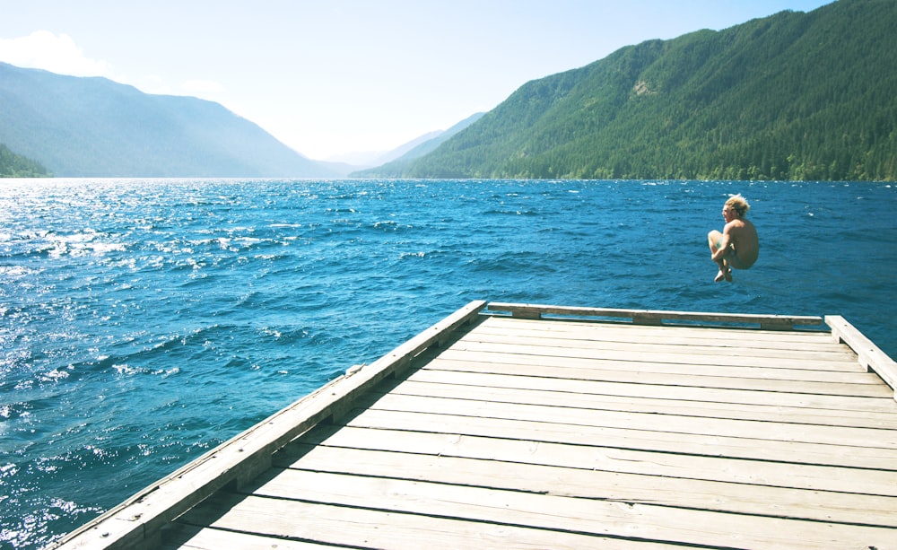 topless person jumping towards water from white wooden dock