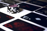 A stack of poker chips with a distinctive black and white pattern is placed on a table covered with several playing cards. The cards are dark with red and white symbols, creating a bold contrast against the lighter surface.