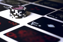 A stack of poker chips with a distinctive black and white pattern is placed on a table covered with several playing cards. The cards are dark with red and white symbols, creating a bold contrast against the lighter surface.