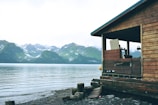A wooden cabin overlooks a tranquil body of water with a backdrop of lush, green mountains and snow-capped peaks. The cabin has a rustic appearance, with weathered wood and a simple, open porch. The shoreline is rocky, partially covered with stones and driftwood.