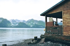 A wooden cabin overlooks a tranquil body of water with a backdrop of lush, green mountains and snow-capped peaks. The cabin has a rustic appearance, with weathered wood and a simple, open porch. The shoreline is rocky, partially covered with stones and driftwood.