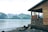 A wooden cabin overlooks a tranquil body of water with a backdrop of lush, green mountains and snow-capped peaks. The cabin has a rustic appearance, with weathered wood and a simple, open porch. The shoreline is rocky, partially covered with stones and driftwood.