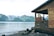 A wooden cabin overlooks a tranquil body of water with a backdrop of lush, green mountains and snow-capped peaks. The cabin has a rustic appearance, with weathered wood and a simple, open porch. The shoreline is rocky, partially covered with stones and driftwood.