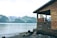 A wooden cabin overlooks a tranquil body of water with a backdrop of lush, green mountains and snow-capped peaks. The cabin has a rustic appearance, with weathered wood and a simple, open porch. The shoreline is rocky, partially covered with stones and driftwood.