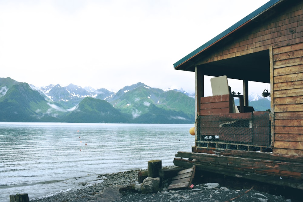 cabina di legno marrone a riva con vista sulla catena montuosa verde attraverso lo specchio d'acqua durante il giorno