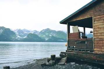 A wooden cabin overlooks a tranquil body of water with a backdrop of lush, green mountains and snow-capped peaks. The cabin has a rustic appearance, with weathered wood and a simple, open porch. The shoreline is rocky, partially covered with stones and driftwood.