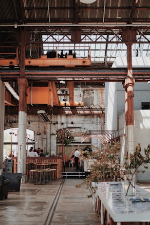An industrial-style interior space with high ceilings and exposed metal beams. The area features rustic and vintage decor with wooden tables and chairs. There are plants adding a touch of greenery, and the lighting is warm and inviting. People are gathered in a communal area, possibly a café or restaurant.