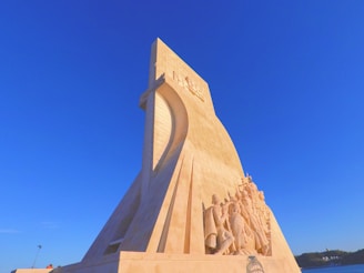 A large, intricately carved stone monument stands against a deep blue sky. It features detailed sculptures of historical figures depicted in flowing robes, positioned as if they are embarking on a journey. The structure has an angular, modernist design with a prominent vertical element and reliefs that create shadows in the afternoon light.