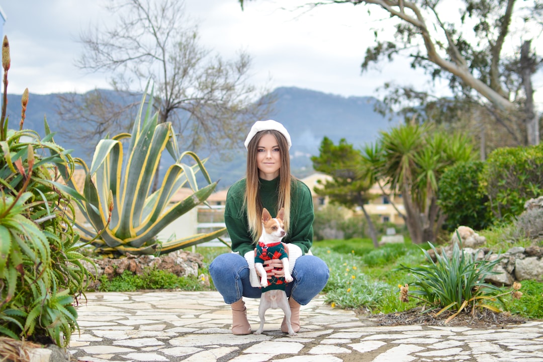 woman wearing green crew-neck long-sleeved shirt holding white and brown dog on pathway beside plants during daytime