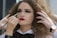 A person with voluminous curly hair receives makeup application. Bold eye makeup and bright red lipstick contrast with their white collared shirt and black bow tie. Red nails match the lipstick, and a watch adorns their wrist.