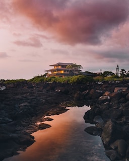 A modern house sits atop a rocky hill overlooking a serene body of water. The sky is filled with dramatic clouds, painted in hues of pink and orange as the sun sets. Lush greenery surrounds the house, contrasting with the dark rocks that line the shore.