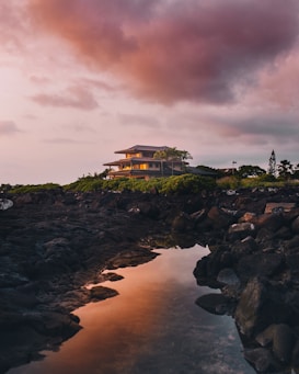 A modern house sits atop a rocky hill overlooking a serene body of water. The sky is filled with dramatic clouds, painted in hues of pink and orange as the sun sets. Lush greenery surrounds the house, contrasting with the dark rocks that line the shore.