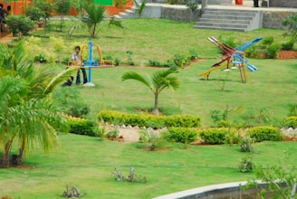 A lush, green park setting with well-maintained lawns and scattered palm trees. In the foreground, there are two colorful playground structures, including a seesaw and a slide. A person is visible near the seesaw. The background shows steps leading to an elevated area with more greenery and a few people walking.