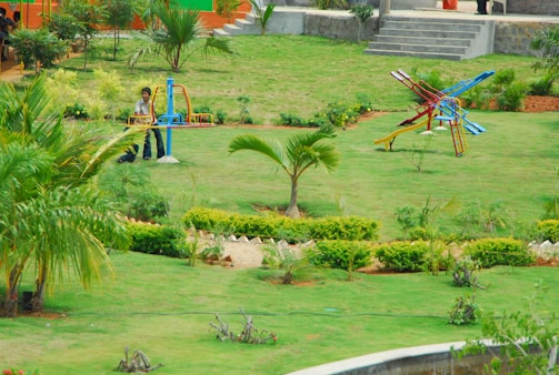 A lush, green park setting with well-maintained lawns and scattered palm trees. In the foreground, there are two colorful playground structures, including a seesaw and a slide. A person is visible near the seesaw. The background shows steps leading to an elevated area with more greenery and a few people walking.