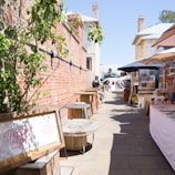 A narrow alleyway lined with red brick walls hosts a market with colorful umbrellas and display tables showcasing various goods. Wooden spool tables are placed along the left side, while people walk and browse the market offerings. A sign reading 'Market Lane' is visible, indicating market days.