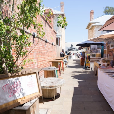 A narrow alleyway lined with red brick walls hosts a market with colorful umbrellas and display tables showcasing various goods. Wooden spool tables are placed along the left side, while people walk and browse the market offerings. A sign reading 'Market Lane' is visible, indicating market days.