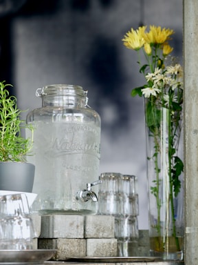 A glass water dispenser with a metal spigot is placed on a stack of concrete blocks. To the left, a small potted plant with green leaves is visible. Several upside-down clear glasses are neatly stacked next to the dispenser. On the right, a tall, clear vase holds yellow and white flowers with green stems.