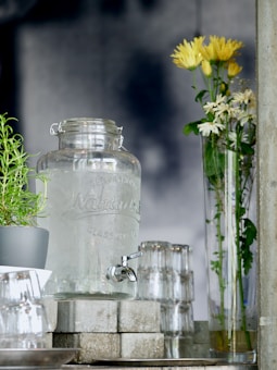 A glass water dispenser with a metal spigot is placed on a stack of concrete blocks. To the left, a small potted plant with green leaves is visible. Several upside-down clear glasses are neatly stacked next to the dispenser. On the right, a tall, clear vase holds yellow and white flowers with green stems.