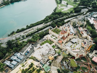 An aerial view of a large amusement park with a prominent castle-like structure featuring red roofs and multiple turrets. Surrounding the castle are various attractions, including roller coasters and several green areas. The amusement park is adjacent to a body of water and bordered by lush greenery and roadways. The scene is bustling with activity and vivid colors.