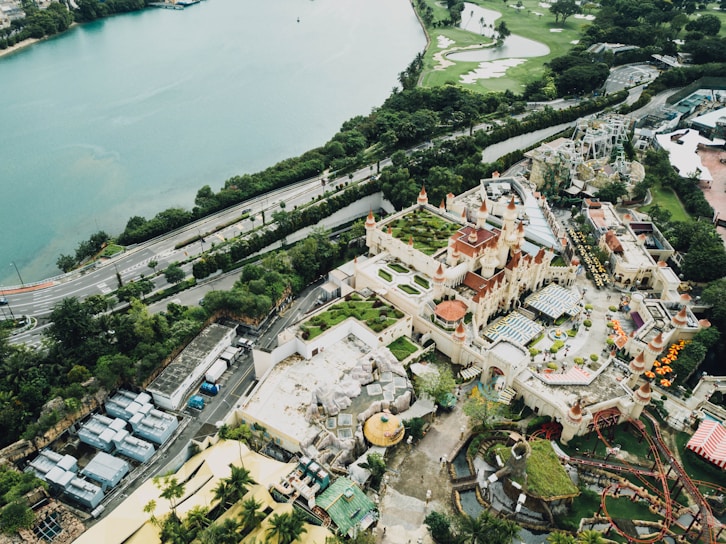 An aerial view of a large amusement park with a prominent castle-like structure featuring red roofs and multiple turrets. Surrounding the castle are various attractions, including roller coasters and several green areas. The amusement park is adjacent to a body of water and bordered by lush greenery and roadways. The scene is bustling with activity and vivid colors.
