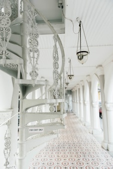 An ornate, white spiral staircase with intricate metalwork is prominently featured. It is set against a long, covered walkway with patterned floor tiles and hanging lamps. An arched column design lines the passageway, and a sign on the staircase reads 'For Staff Only'.
