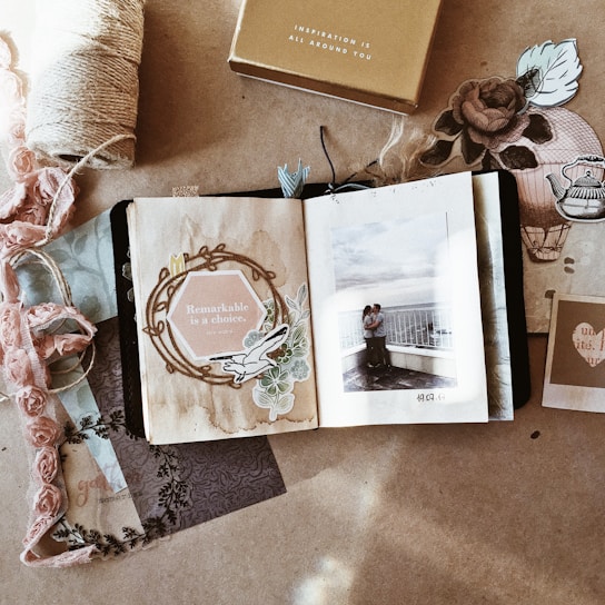 A scrapbook-style composition with an open book displaying a photograph of a couple embracing on a balcony. The left page reads 'Remarkable is a choice' surrounded by floral and vintage elements. Various decorative papers, fabric, and a spool of twine surround the book, creating a cozy and nostalgic atmosphere.