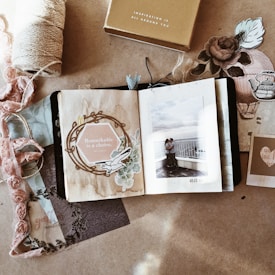 A scrapbook-style composition with an open book displaying a photograph of a couple embracing on a balcony. The left page reads 'Remarkable is a choice' surrounded by floral and vintage elements. Various decorative papers, fabric, and a spool of twine surround the book, creating a cozy and nostalgic atmosphere.