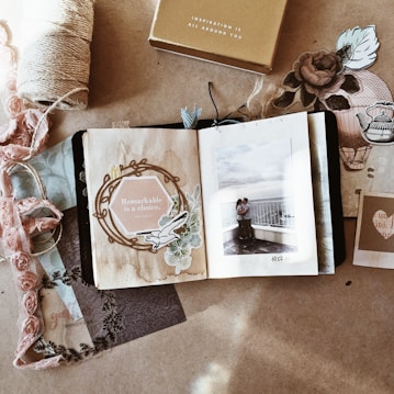A scrapbook-style composition with an open book displaying a photograph of a couple embracing on a balcony. The left page reads 'Remarkable is a choice' surrounded by floral and vintage elements. Various decorative papers, fabric, and a spool of twine surround the book, creating a cozy and nostalgic atmosphere.