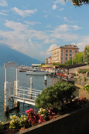 A serene lakeside scene with clear blue skies, featuring a marina with a dock and several boats moored. A row of vibrant flowers and lush greenery lines the waterfront. In the background, there is an elegant, multi-story building with large windows and a visible sign. The surrounding mountains create a calm and picturesque backdrop.