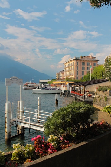 A serene lakeside scene with clear blue skies, featuring a marina with a dock and several boats moored. A row of vibrant flowers and lush greenery lines the waterfront. In the background, there is an elegant, multi-story building with large windows and a visible sign. The surrounding mountains create a calm and picturesque backdrop.
