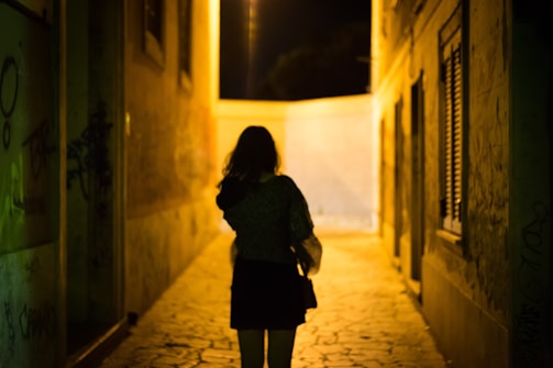 A lone figure in nocturna gear walking through a graffiti-covered alley illuminated by street lamps.