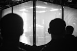 Silhouettes of two people are seen facing a large glass enclosure. Inside the enclosure, bright lights illuminate a stage setup with electronic equipment, creating a high-contrast scene.