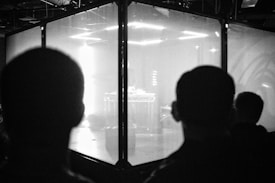 Silhouettes of two people are seen facing a large glass enclosure. Inside the enclosure, bright lights illuminate a stage setup with electronic equipment, creating a high-contrast scene.