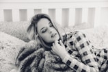 A person with closed eyes rests on a plush bed with textured pillows, wearing a patterned outfit. The setting has a cozy and intimate feel, enhanced by the soft focus and monochrome color scheme.