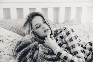 A person with closed eyes rests on a plush bed with textured pillows, wearing a patterned outfit. The setting has a cozy and intimate feel, enhanced by the soft focus and monochrome color scheme.