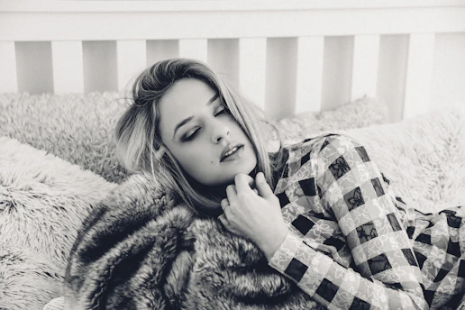 A person with closed eyes rests on a plush bed with textured pillows, wearing a patterned outfit. The setting has a cozy and intimate feel, enhanced by the soft focus and monochrome color scheme.
