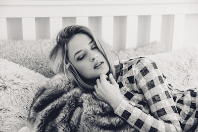 A person with closed eyes rests on a plush bed with textured pillows, wearing a patterned outfit. The setting has a cozy and intimate feel, enhanced by the soft focus and monochrome color scheme.