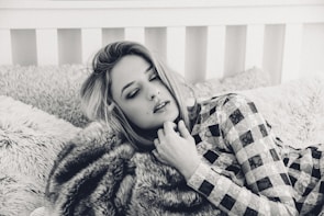 A person with closed eyes rests on a plush bed with textured pillows, wearing a patterned outfit. The setting has a cozy and intimate feel, enhanced by the soft focus and monochrome color scheme.