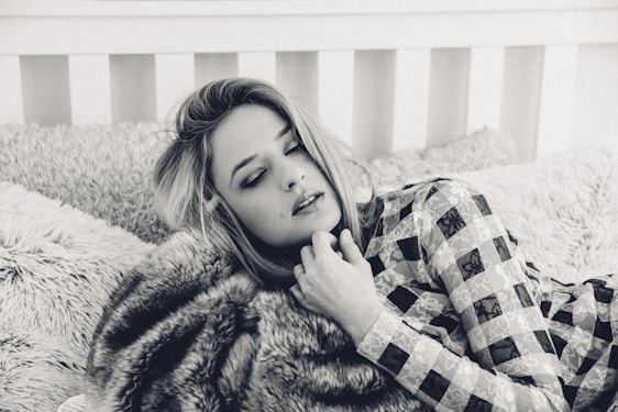 A person with closed eyes rests on a plush bed with textured pillows, wearing a patterned outfit. The setting has a cozy and intimate feel, enhanced by the soft focus and monochrome color scheme.