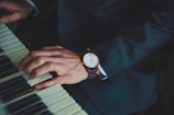 A close-up of a person wearing a dark suit, playing a piano. The person's left hand is visible, displaying a wristwatch with a brown leather band. The piano keys are partially in view, highlighting both black and white keys.
