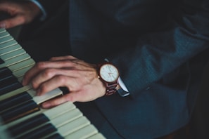 A close-up of a person wearing a dark suit, playing a piano. The person's left hand is visible, displaying a wristwatch with a brown leather band. The piano keys are partially in view, highlighting both black and white keys.