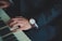 A close-up of a person wearing a dark suit, playing a piano. The person's left hand is visible, displaying a wristwatch with a brown leather band. The piano keys are partially in view, highlighting both black and white keys.